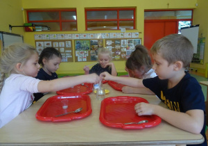 Dzieci siedzą przy stoliku, w ręce trzymają pipetki, które napełniają wodą i przelewają do pojemników z drożdżami..